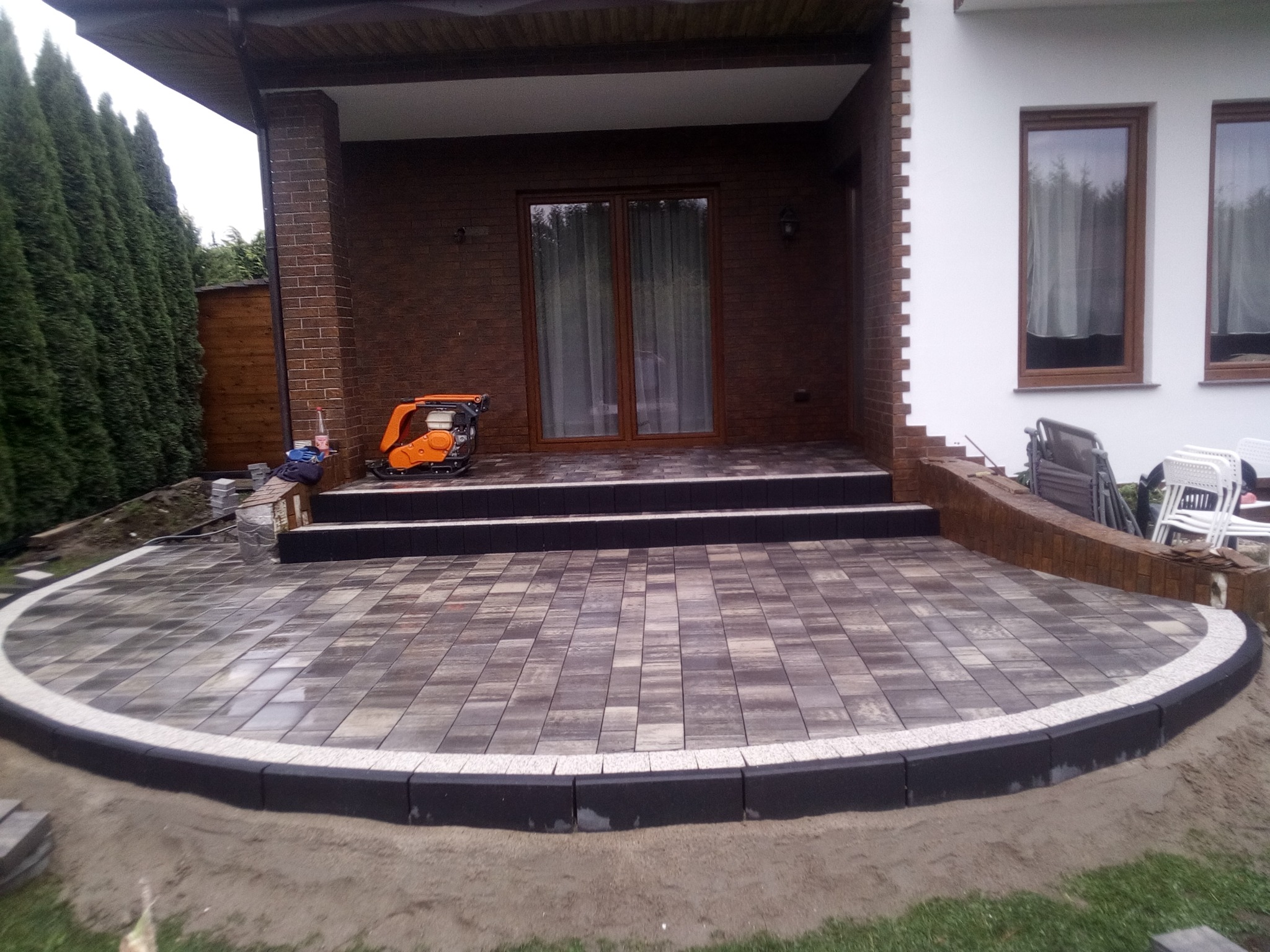 Półokrągły taras z kostki brukowej w odcieniach szarości, obrzeża wykończone czarnym krawężnikiem z jasną listwą, w tle budynek z ceglaną elewacją i widoczną zagęszczarką.