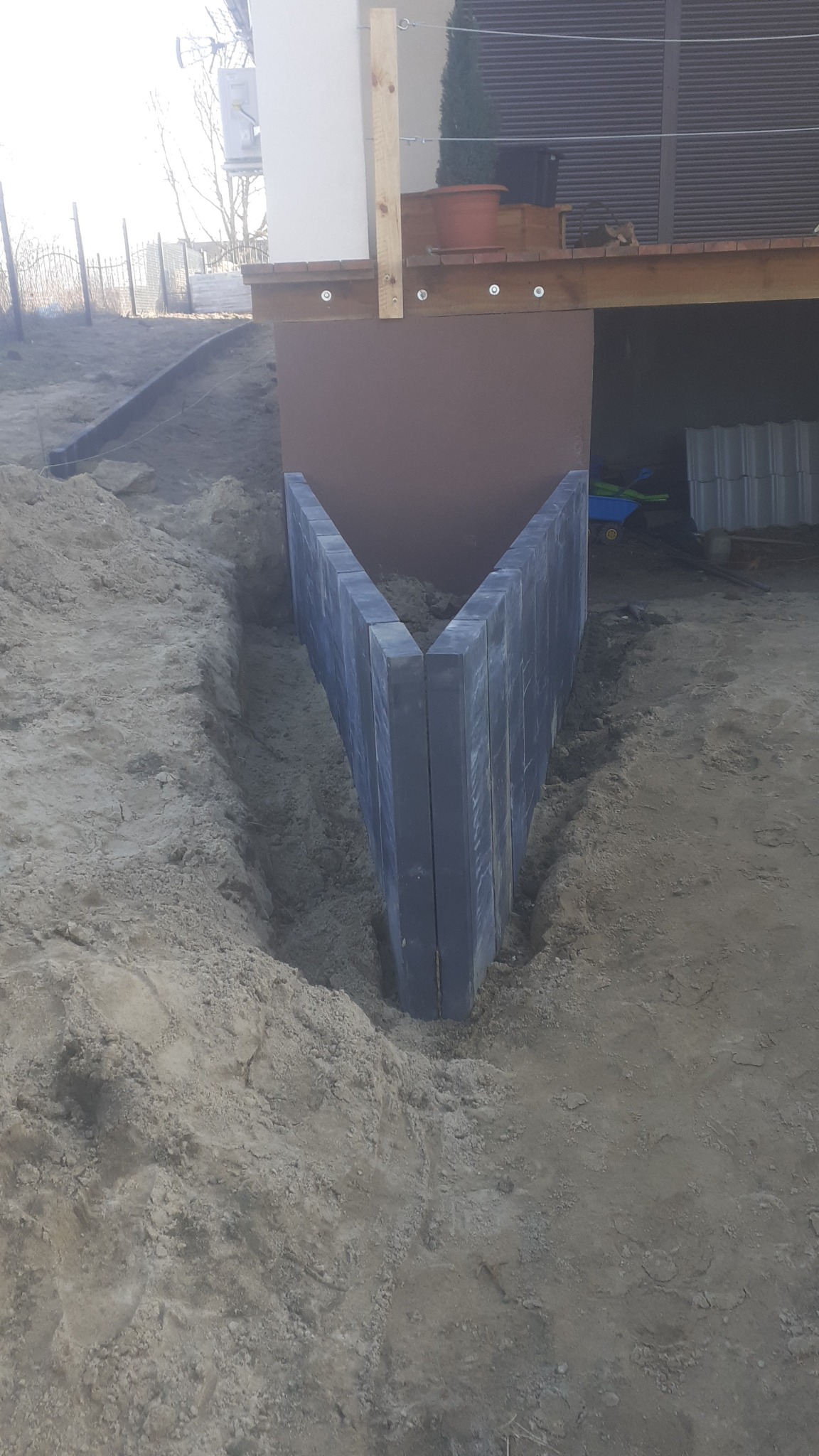 Wkopane w ziemię, pionowo ustawione, szare, betonowe elementy tworzą kąt prosty, prawdopodobnie przygotowanie pod budowę opaski wokół budynku.