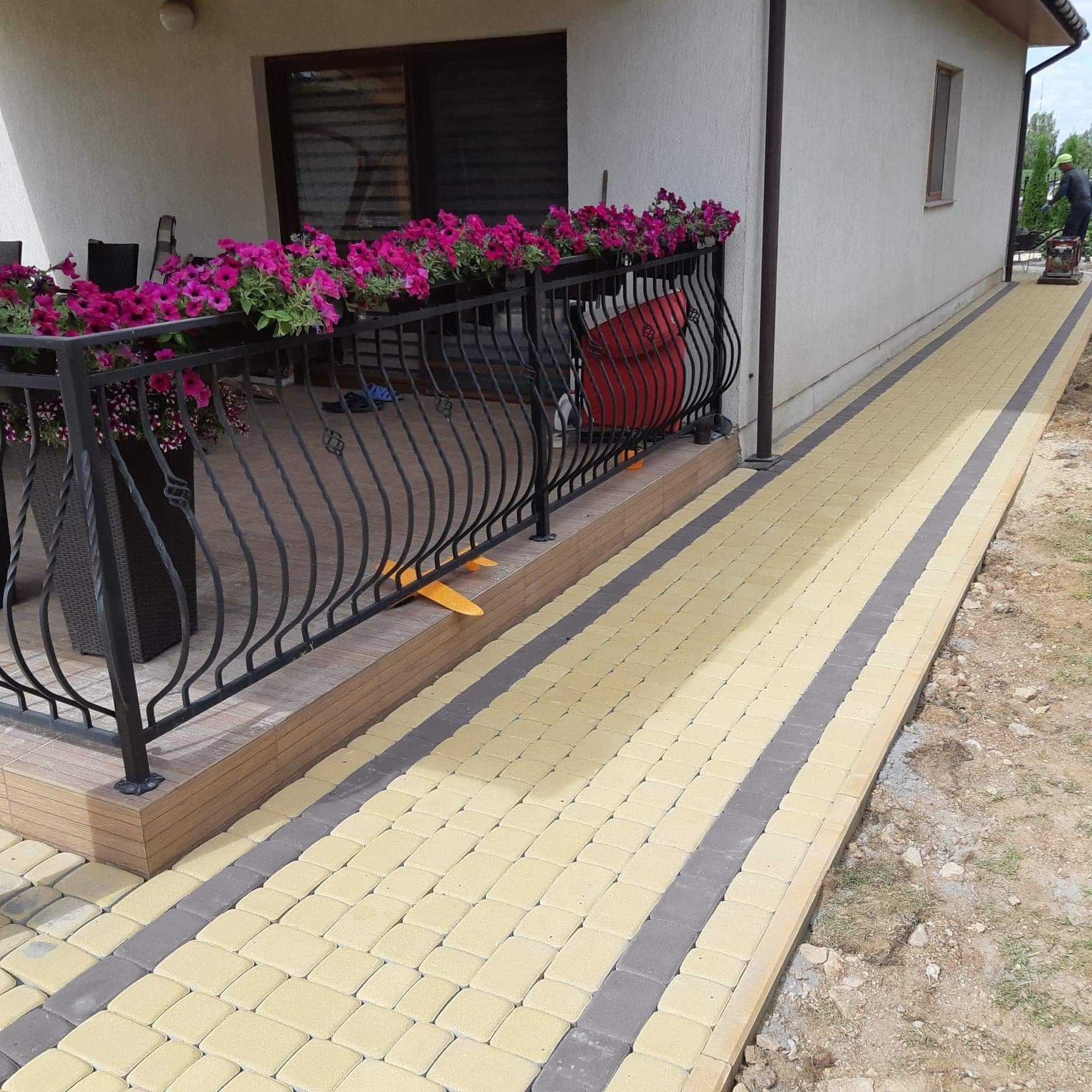 Ułożona kostka brukowa w kolorze żółtym i szarym prowadząca od tarasu z czarną balustradą i różowymi kwiatami, pracownik w tle zagęszcza podłoże.