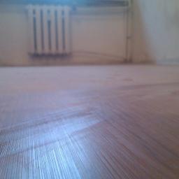 Fin end - Ukończona drewniana podłoga w pokoju z białym grzejnikiem na ścianie. Widok z niskiej perspektywy ukazuje detale wykończenia.