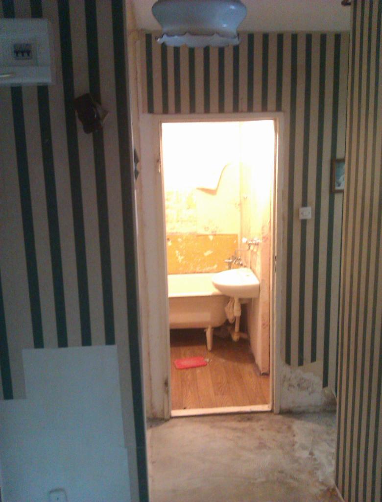 Łazienka w remoncie: widok na wnętrze z wanną na nóżkach i umywalką, przez futrynę drzwiową. Tapeta w pionowe pasy i fragment posadzki w kadrze.