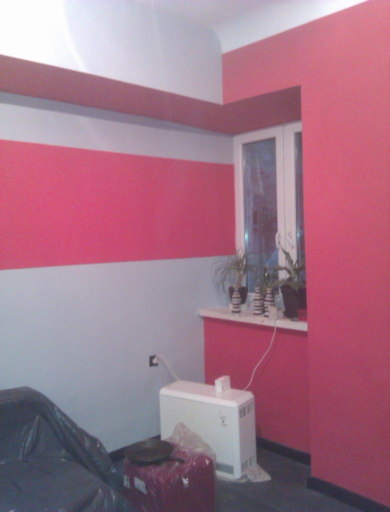 Ściana po malowaniu w paski: różowy i szary. Widoczny grzejnik elektryczny, okno z kwiatami i zabezpieczone folią meble. Akcent kolorystyczny na suficie.
