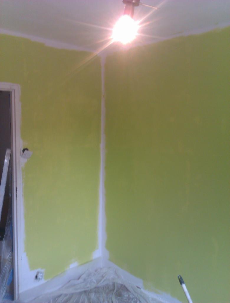 Ściana pomalowana na jasnozielony kolor, widoczne białe obramowania i zabezpieczona folią podłoga. W rogu pokoju widoczny fragment drabiny.