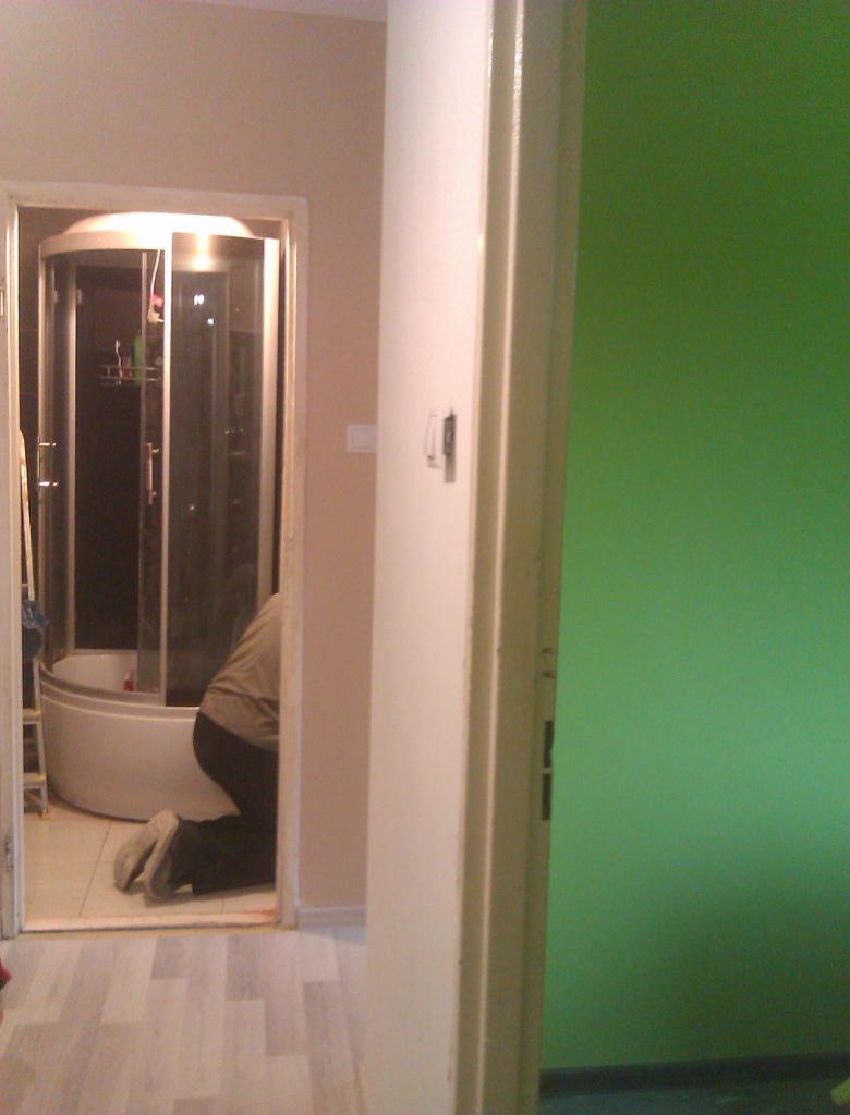 Instalacja kabiny prysznicowej w łazience. Widoczna osoba na kolanach, kabina z hydromasażem i fragment zielonej ściany w tle.