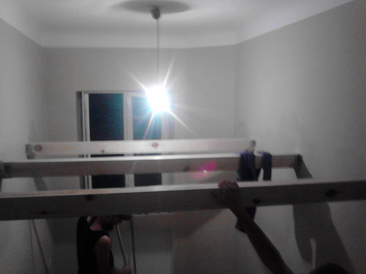 Pokój w trakcie malowania: drabina oparta o ścianę, widoczne ręce, okno w tle. Lampa sufitowa z intensywnym światłem. Etap prac wykończeniowych.