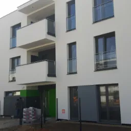 Nowoczesny budynek wielorodzinny z białymi elewacjami i szklanymi balustradami balkonów, z widocznymi workami materiałów budowlanych przed wejściem i pracującym mężczyzną w tle.