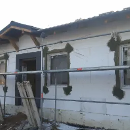 Budowa domu z białych bloczków, widoczne rusztowanie, przygotowane otwory okienne z czarną ramą, częściowo ocieplone ściany, niezabezpieczone okna, drewniana konstrukcja dachu.