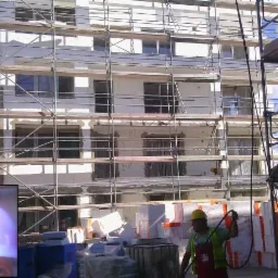 Ocieplanie elewacji budynku wielorodzinnego ze stalowymi rusztowaniami, widoczny pracownik w kasku i kamizelce odblaskowej oraz materiały izolacyjne.