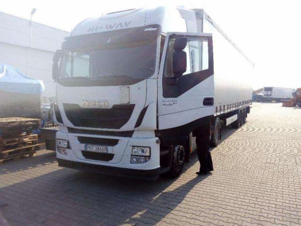 Biała ciężarówka Iveco Hi-Way z otwartymi drzwiami kabiny, zaparkowana na brukowanym placu, z paletami i przykrytym ładunkiem w tle.