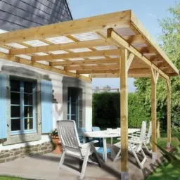 Drewniana pergola z przezroczystym zadaszeniem, przylegająca do białego domu z błękitnymi okiennicami. Na tarasie białe meble ogrodowe.