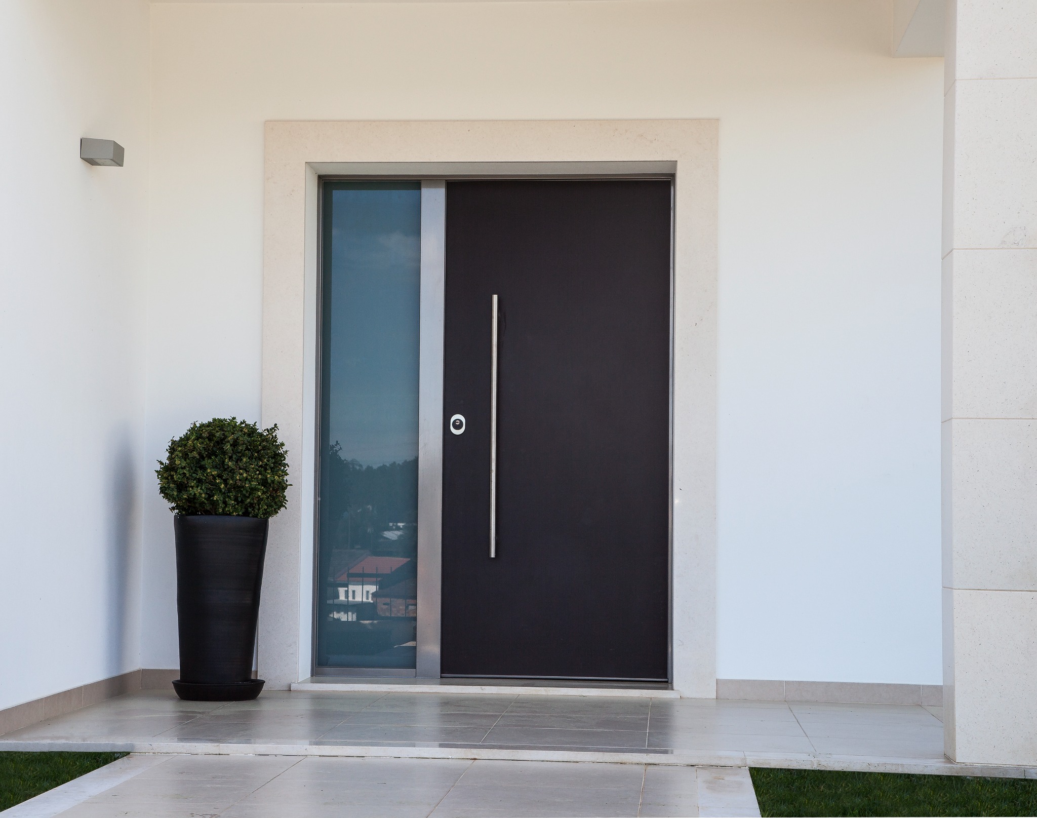 Nowoczesne drzwi wejściowe w kolorze antracytowym z długim, pionowym uchwytem i szybą, umieszczone w jasnej, kamiennej ramie. Obok stoi wysoka, czarna donica z zieloną rośliną.