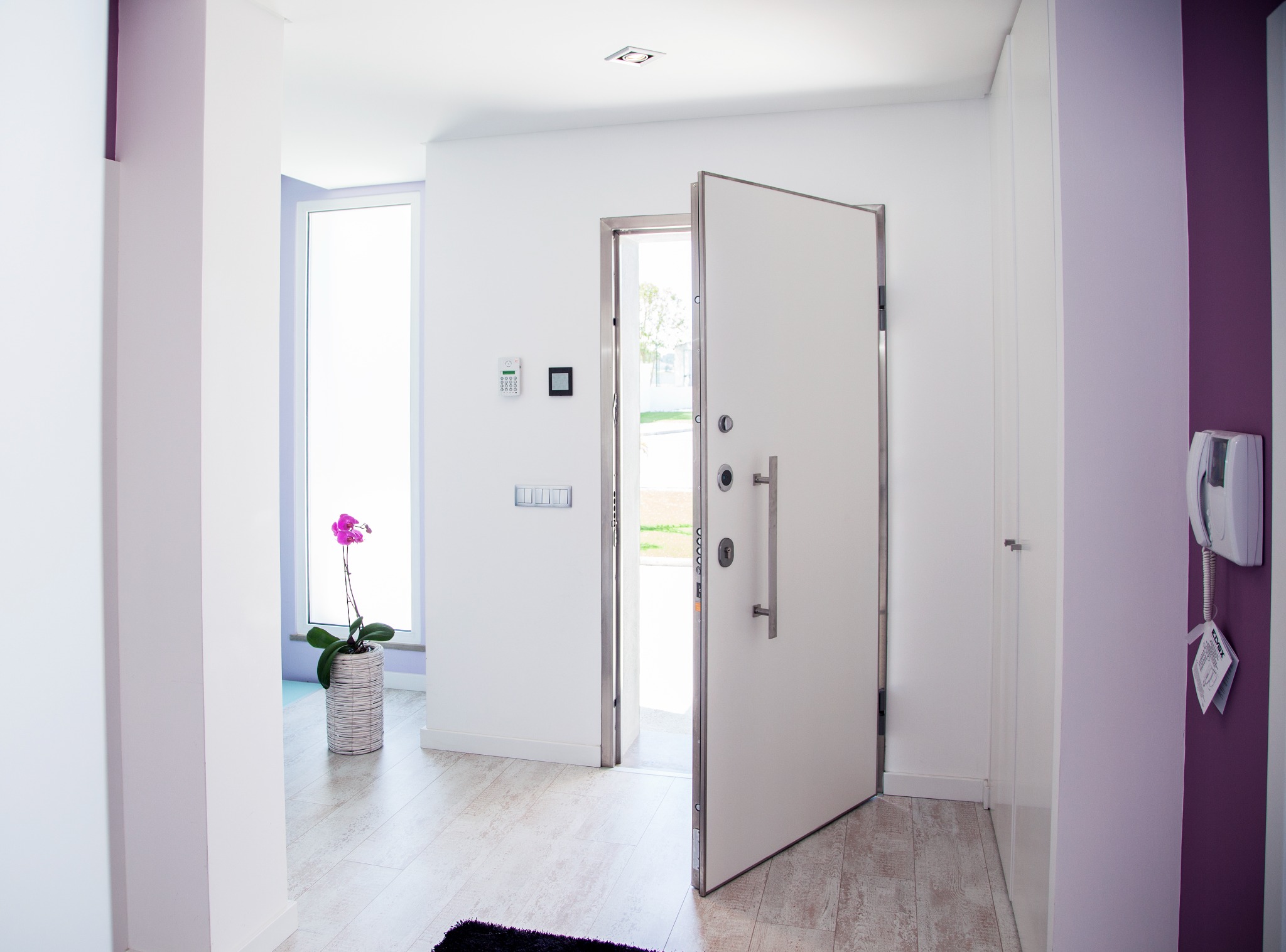 Otwarta, biała, nowoczesna  z metalowym obramowaniem. Widoczny zamek, wizjer i duży uchwyt. W tle jasne wnętrze z fioletowymi akcentami i kwiatem.