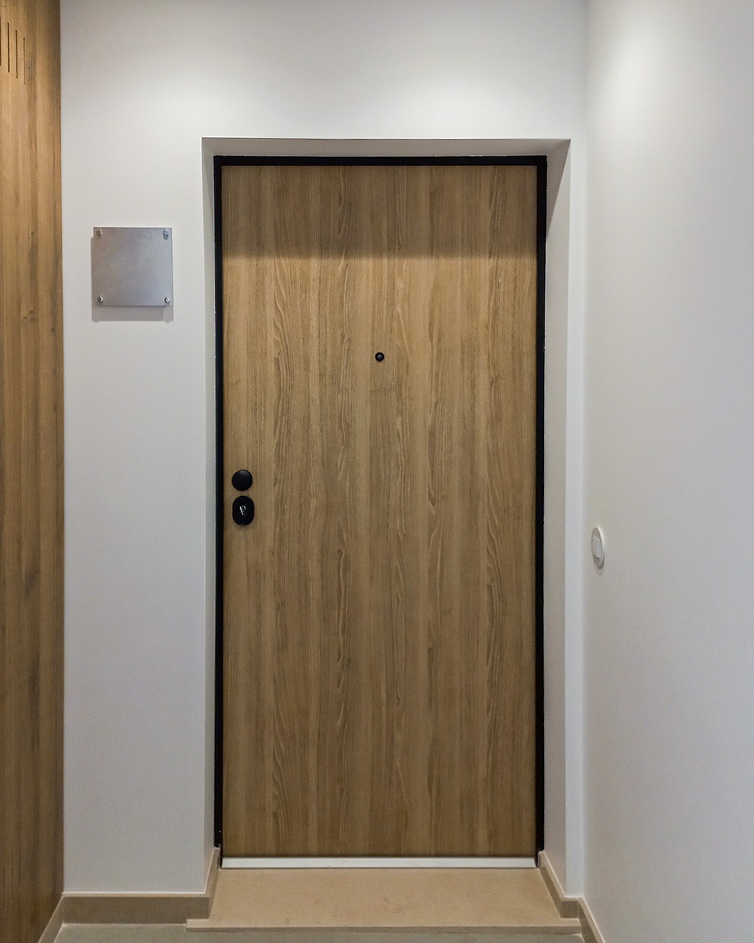 Nowoczesne drzwi wejściowe z wizjerem, w kolorze jasnego drewna, z czarną ramą, zamontowane w jasnym korytarzu z płytką tabliczką informacyjną po lewej stronie.