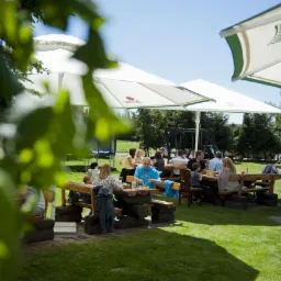 Grupa osób siedzi przy drewnianych stołach na trawniku pod białymi parasolami w plenerze, widoczny plac zabaw w tle.