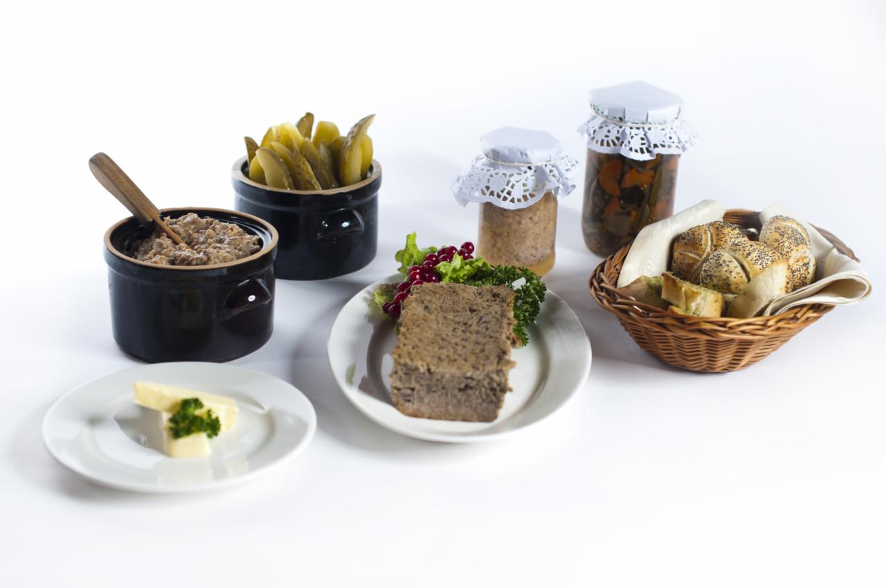 Szeroki kadr przedstawia różnorodne dania: pasztet w słoikach z koronkowymi nakładkami, domowa pasta w ciemnym naczyniu z drewnianą łyżką, ogórki kiszone w kamionkowym garnku, pieczywo w wiklinowym...