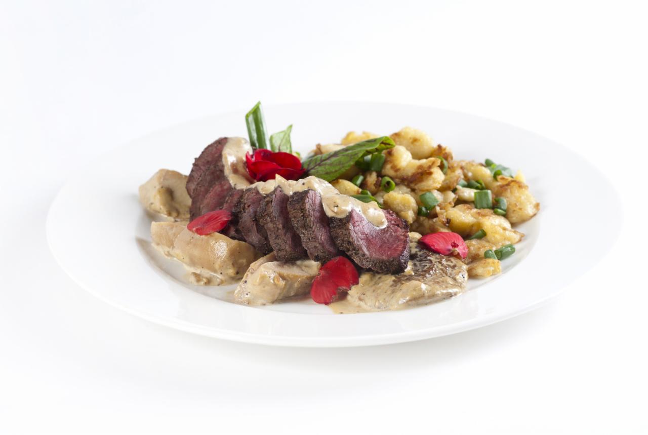 Elegancko podane plastry pieczonej polędwicy z sosem, ziemniaczanymi kluskami i dekoracją z jadalnych kwiatów na białym talerzu.