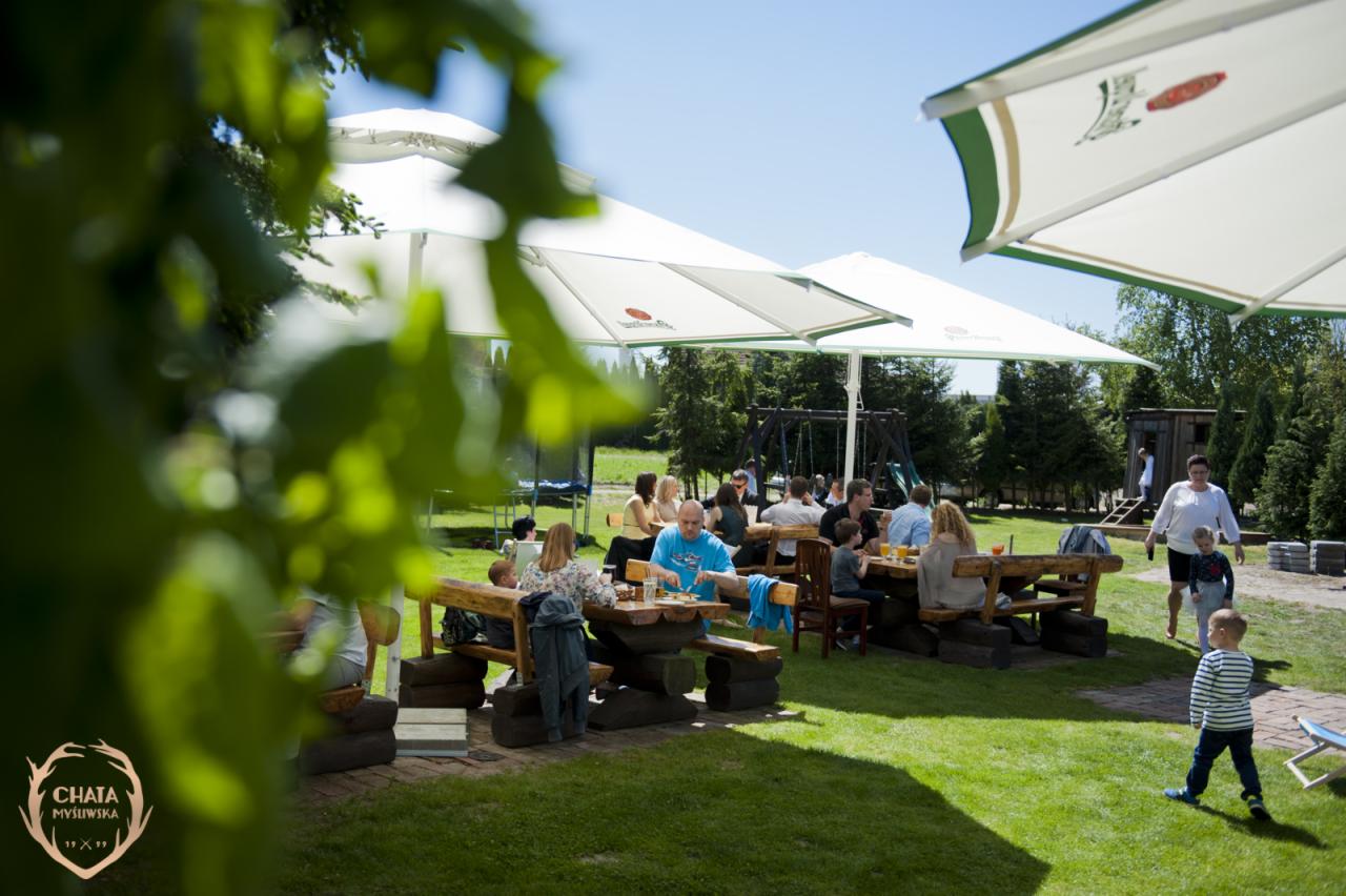 Grupa osób siedzi przy drewnianych stołach na trawniku pod białymi parasolami w plenerze, widoczny plac zabaw w tle.