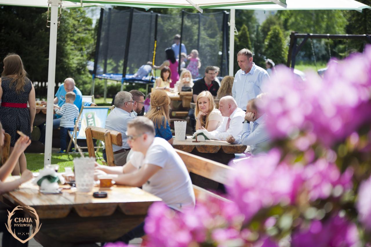 Zewnętrzne przyjęcie z drewnianymi stołami i krzesłami, goście siedzą i rozmawiają, w tle trampolina i huśtawka, w pierwszym planie rozmyte różowe kwiaty.