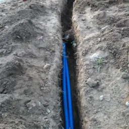 Dwa niebieskie kable karbowane ułożone w wykopie ziemnym, widoczne ślady pracy ziemnej i narzędzia w tle.