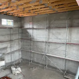 Wnętrze budynku w trakcie tynkowania maszynowego ścian, widoczne rusztowania, surowy strop drewniany, okno oraz worki z materiałami budowlanymi na podłodze.