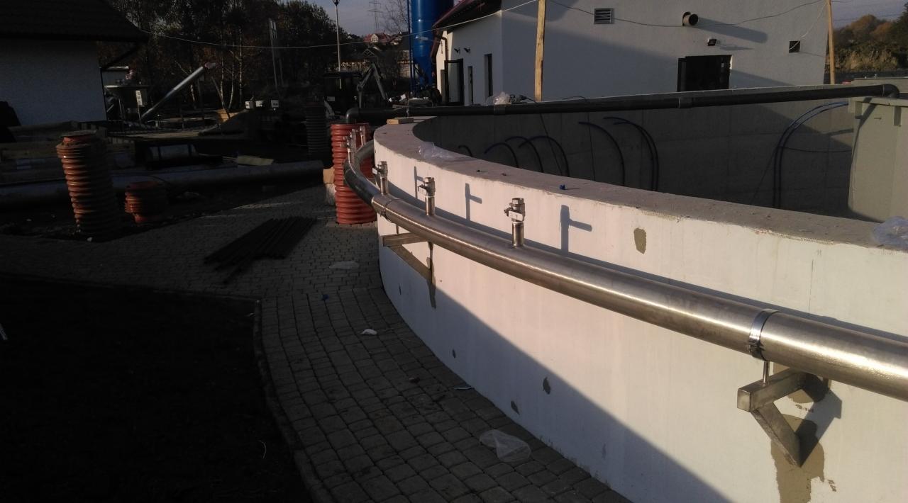 Zakrzywiona balustrada ze stali nierdzewnej, zamontowana na betonowym murku, widoczna kostka brukowa i elementy placu budowy w tle.