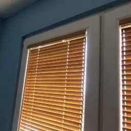 Zamknięte żaluzje drewniane w oknie z białymi ramami na tle niebieskiej ściany, widoczne pod kątem.