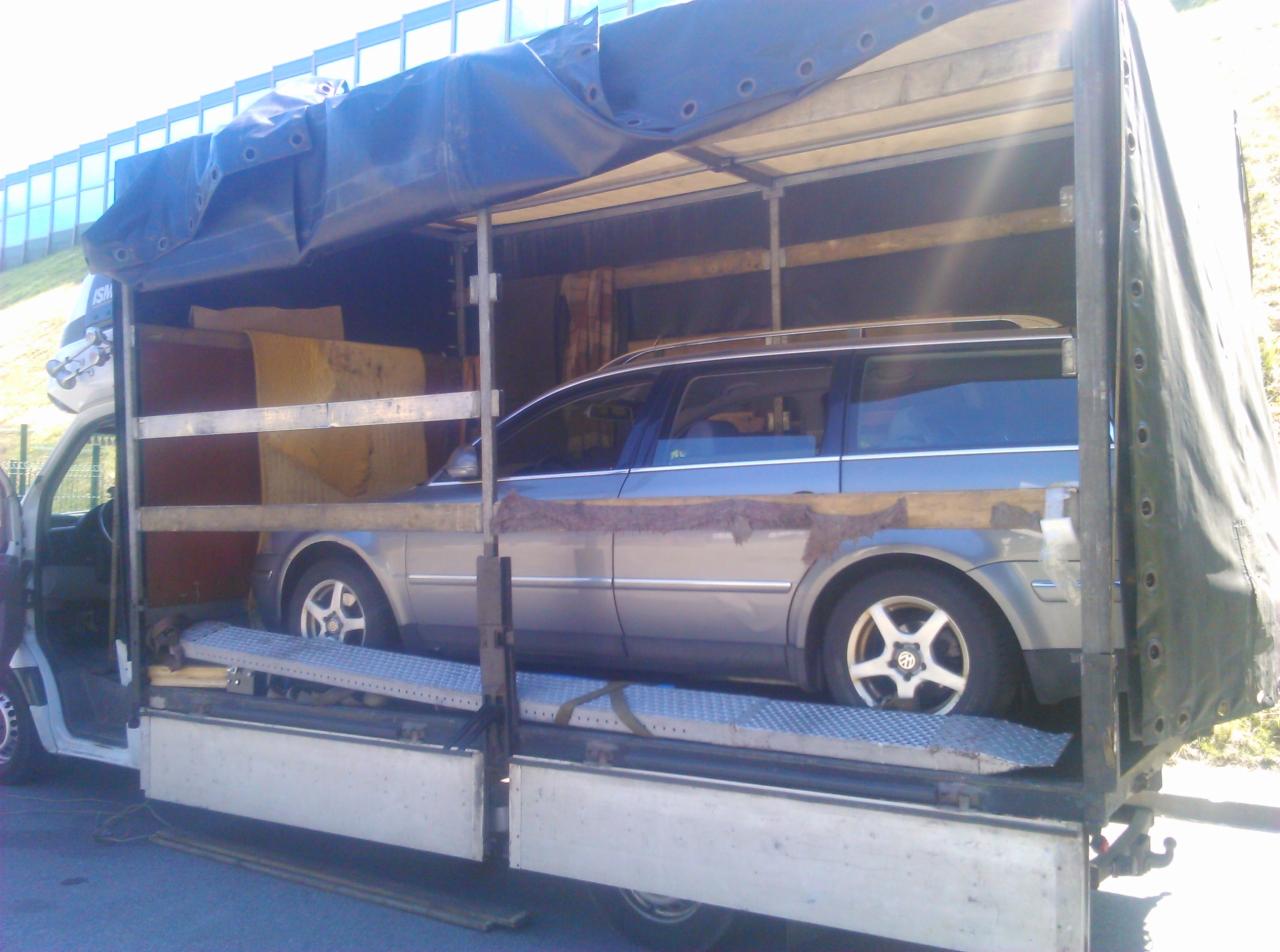 Szary samochód kombi zabezpieczony drewnianymi belkami wewnątrz ciężarówki z plandeką, widoczna rampa załadunkowa i elementy mocujące ładunek.