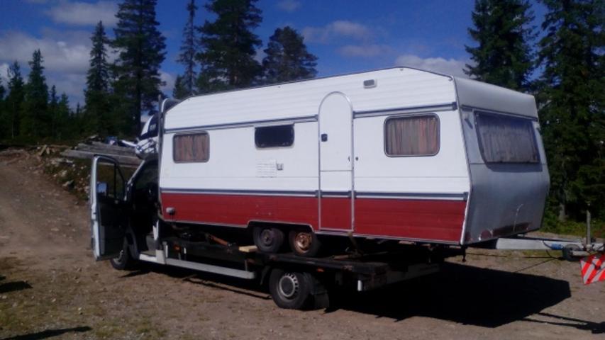 Laweta z przyczepą kempingową na tle leśnym. Otwarta kabina lawety. Przyczepa biało-czerwona, zabezpieczona na lawecie. Widoczne drzewa i teren leśny w tle.