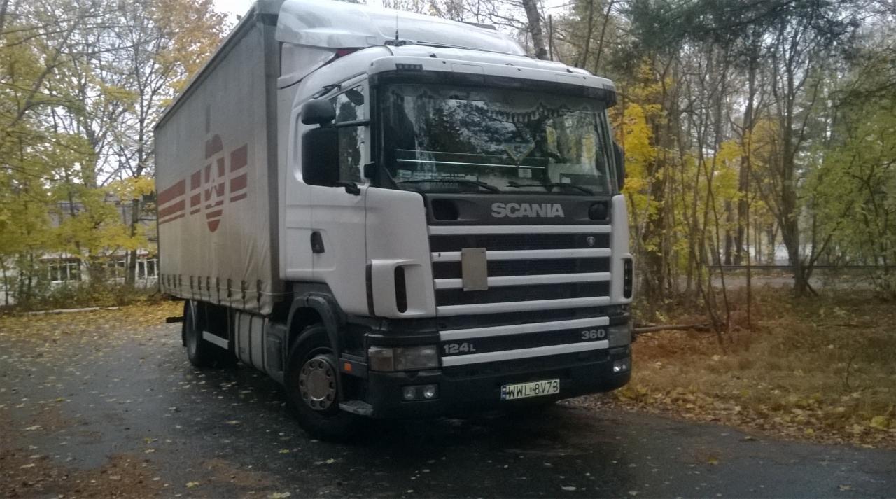 Ciężarówka Scania 124L 360 z naczepą, zaparkowana na mokrym asfalcie w jesiennej scenerii, z widocznym logo firmy transportowej na plandece naczepy.