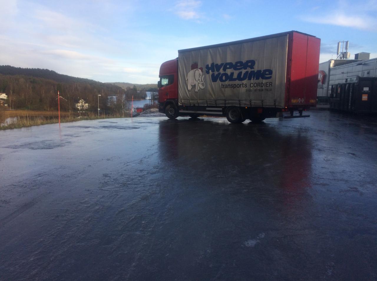 Czerwona ciężarówka Hyper Volume zaparkowana na mokrym placu z widokiem na jezioro i las w tle.