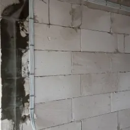 Białe przewody elektryczne ułożone w korytkach na surowej ścianie z betonu komórkowego, zabezpieczone uchwytami, widoczne kable wychodzące ze ściany.