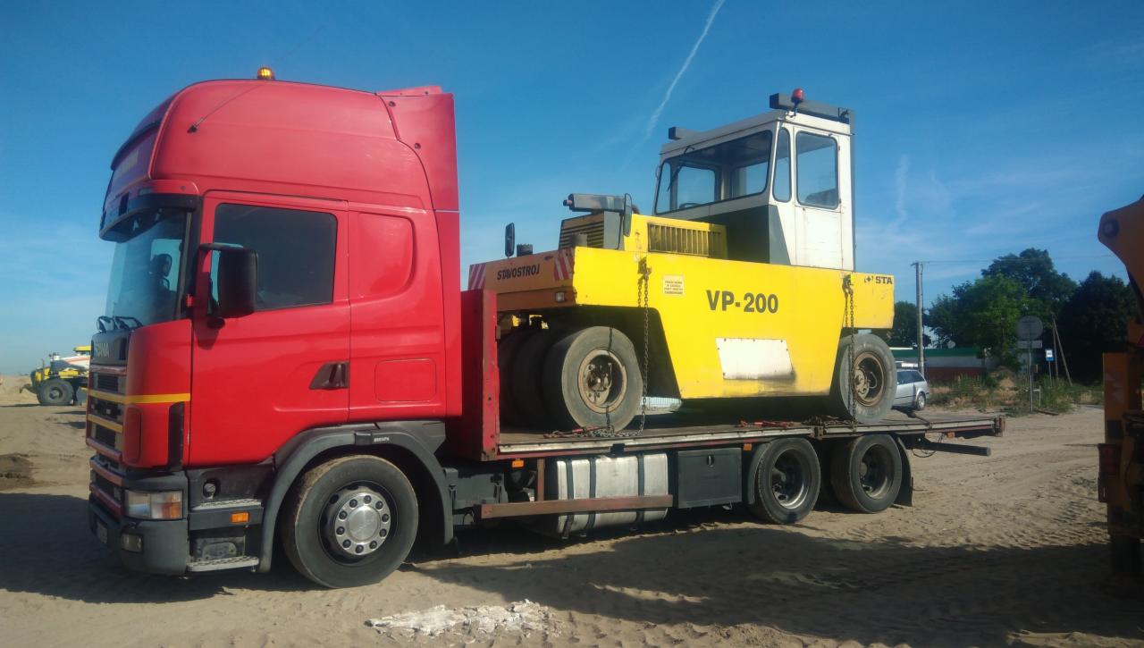 Czerwona ciężarówka transportuje żółty walec drogowy VP-200 na niskopodwoziowej platformie, widok z boku na placu budowy.