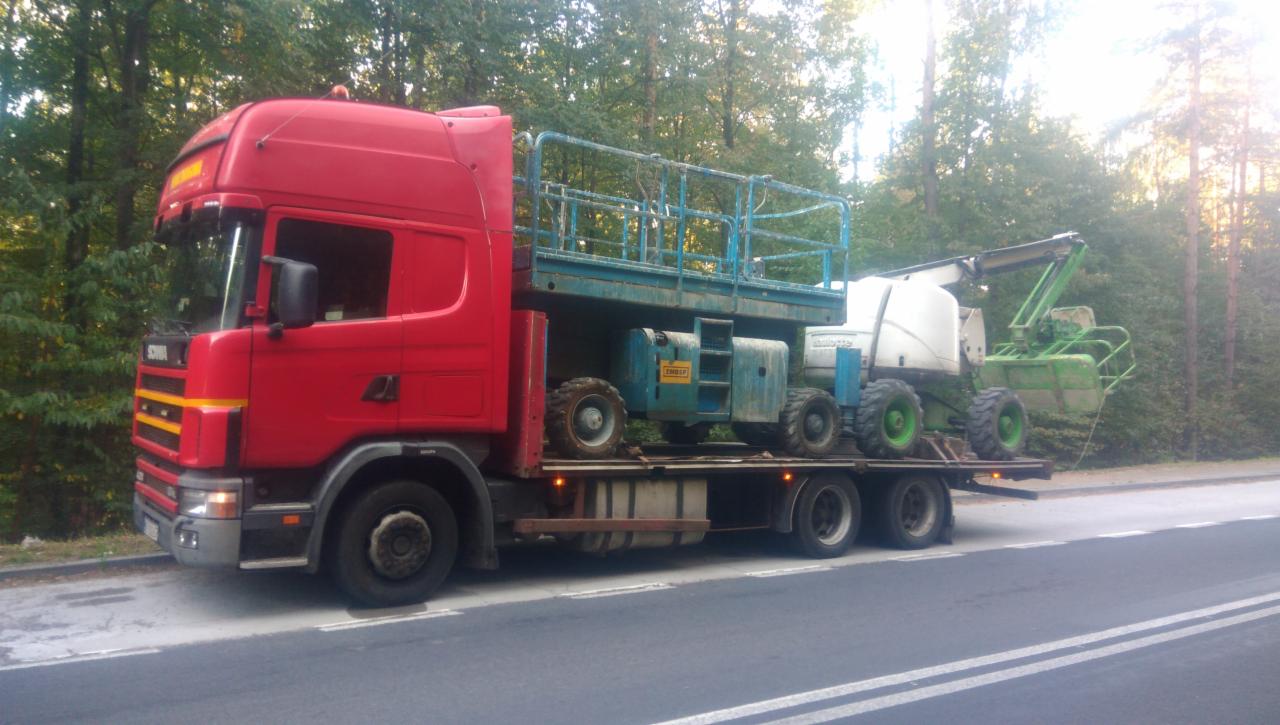 Czerwona ciężarówka Scania przewozi na platformie podnośniki koszowe: niebieski Genie i zielono-biały JLG, na tle lasu.