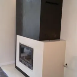 Nowoczesny kominek z czarną obudową i białym frontem, widoczny fragment drabiny i akcesoria montażowe. Ujęcie z boku, podkreślające minimalistyczny design.