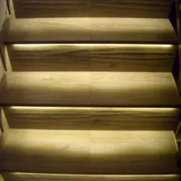 Nowoczesne schody z drewnianymi stopniami, podświetlane delikatnym, ciepłym światłem LED, tworzącym nastrojowy efekt. Widok z dołu, ukazujący detale wykończenia.