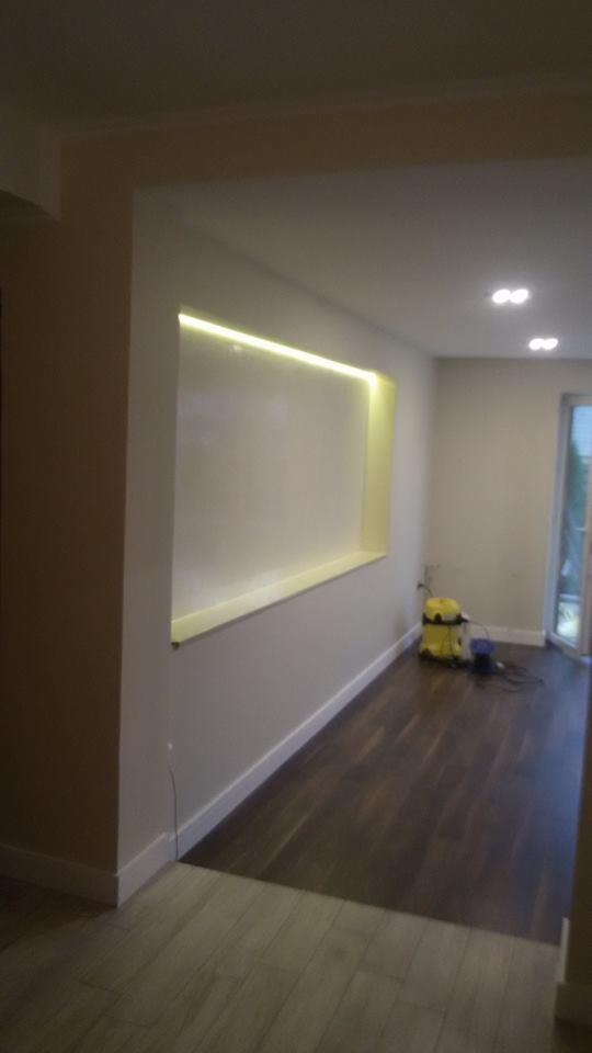 Wnęka ścienna z podświetleniem LED w wykończonym wnętrzu. Widoczna część podłogi i odkurzacz. Minimalistyczny design. Oświetlenie punktowe na suficie.