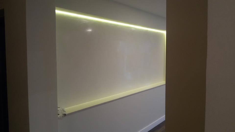 Ściana z wnęką oświetloną listwą LED, widoczna zza filaru. Minimalistyczny design, ciepłe światło podkreśla gładką powierzchnię ściany.