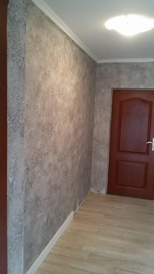 Wnętrze po remoncie: ściany z dekoracyjną fakturą w odcieniach szarości, drewnopodobna podłoga i ciemne drzwi z listwami przypodłogowymi. Oświetlenie sufitowe.