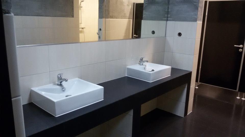 Dwie białe, kwadratowe umywalki z bateriami na czarnym blacie w łazience wyłożonej białymi płytkami. W tle lustro i czarne drzwi.