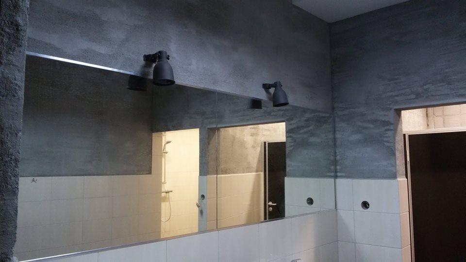 Wykończona łazienka z dużym lustrem i surowymi szarymi ścianami. Widoczne białe płytki, czarne lampy nad lustrem i fragment kabiny prysznicowej w odbiciu.