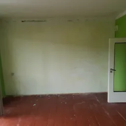 Pokój przed remontem: ściany w odcieniach zieleni i beżu z widocznymi śladami wilgoci przy suficie, drewniana podłoga z resztkami gruzu, otwarte drzwi balkonowe i wewnętrzne.