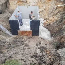 Dwaj pracownicy w pomarańczowych rękawicach uszczelniają betonowy zbiornik w wykopie ziemnym, widoczny metalowy mostek i drabina oparta o ścianę wykopu.