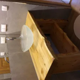 Szklana umywalka na blacie z surowego drewna z widocznymi słojami i korą, zamontowana w łazience z szarymi płytkami i drewnianymi drzwiami.