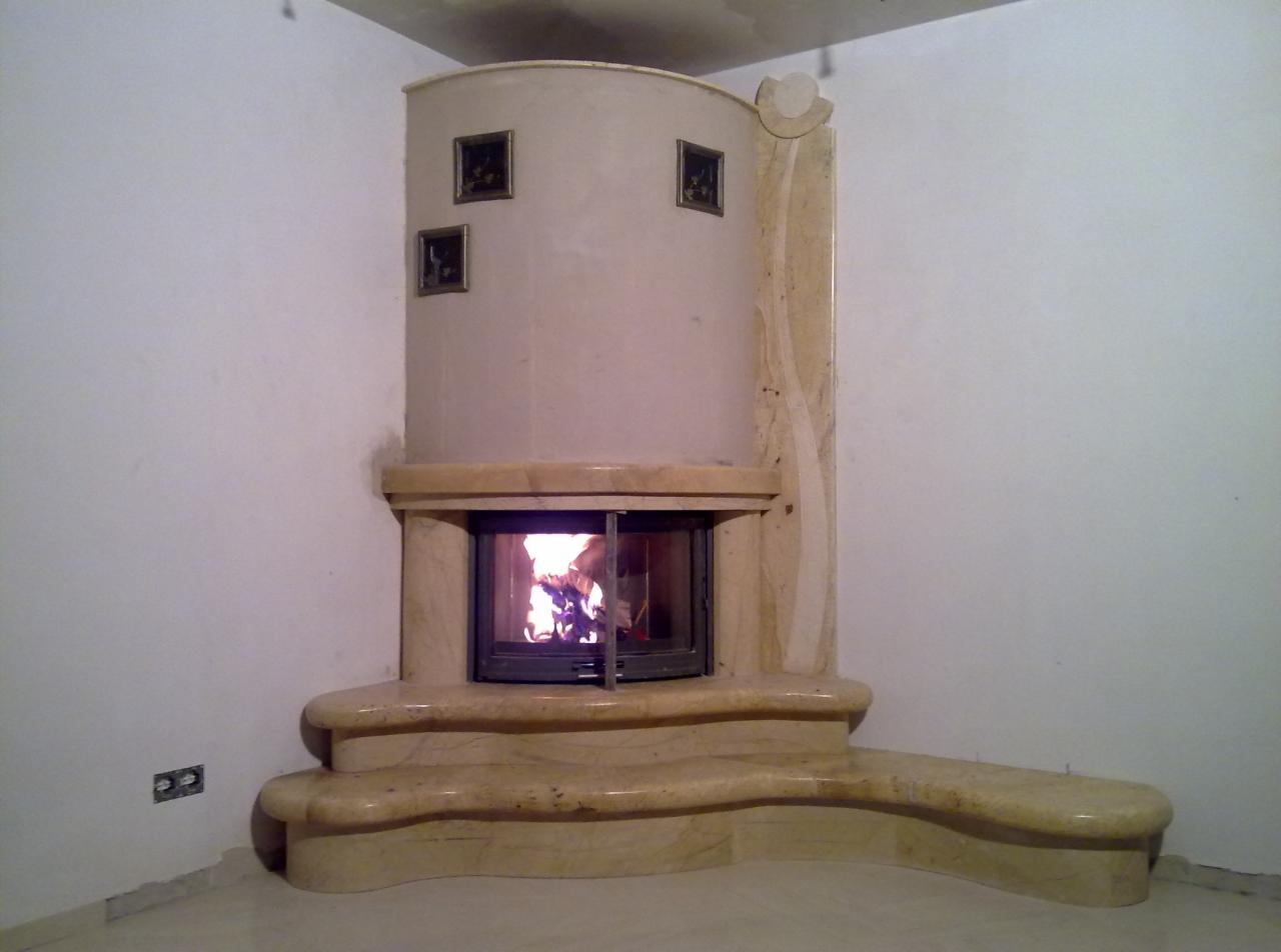 Narożny kominek obudowany jasnym kamieniem z otwartymi drzwiczkami, wewnątrz widoczny płomień, na obudowie trzy małe obrazki w ramkach.