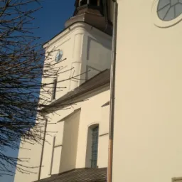 Jasna elewacja kościoła z wieżą i zegarem, widok z dołu, z fragmentem drzewa po lewej. Ciemne okna i drzwi kontrastują z fasadą. Miedziane rynny.