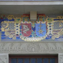 Mozaika heraldyczna nad wejściem do budynku, zdobiona kartuszami z gryfem i krzyżem na gwieździe, otoczona ornamentami i datą. Detal architektoniczny.