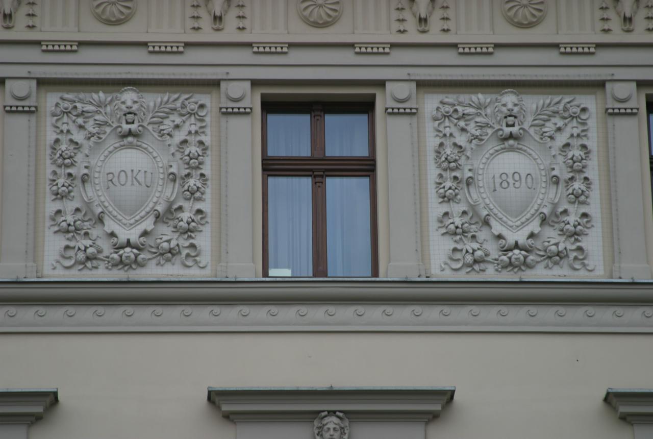 Elewacja kamienicy z dekoracyjnymi płaskorzeźbami, w tym herbami z datami. Okno z drewnianą ramą. Kremowa kolorystyka budynku, detale architektoniczne.