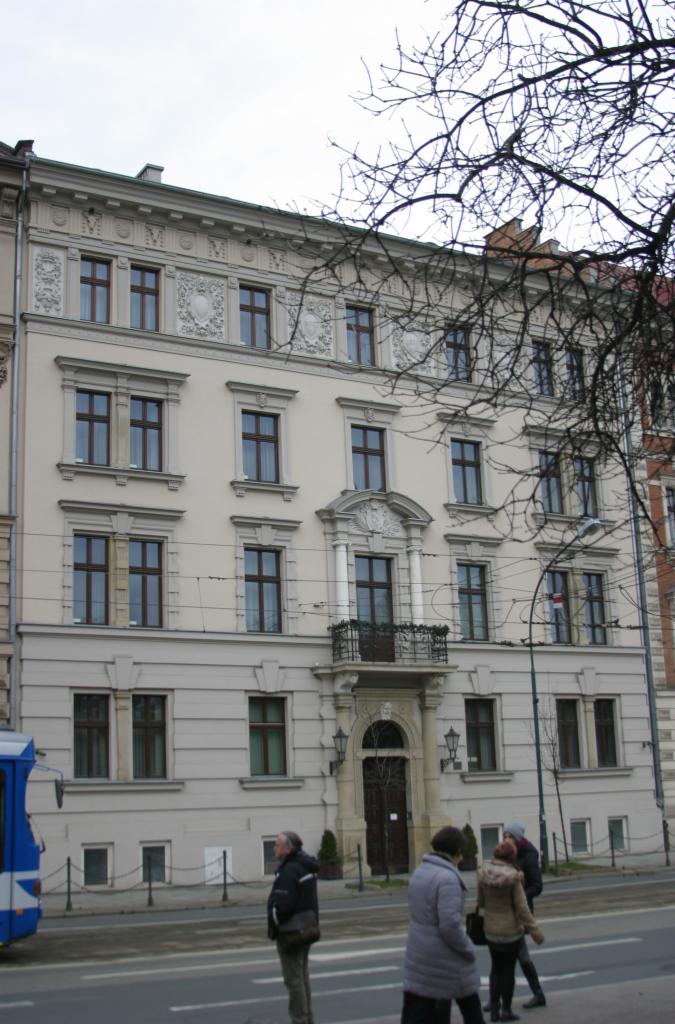 Elewacja kamienicy z balkonem i dekoracyjnymi detalami architektonicznymi. Widoczni przechodnie i fragment tramwaju. Budynek w jasnej kolorystyce.