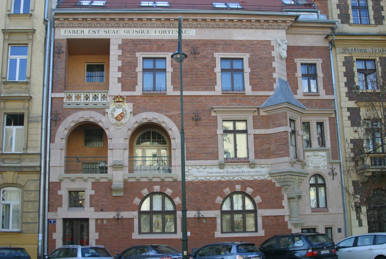 Kamienica z czerwonej cegły z dekoracyjnymi elementami architektonicznymi, balkonami i inskrypcjami. Widoczne detale fasady, okna i elementy zdobnicze.