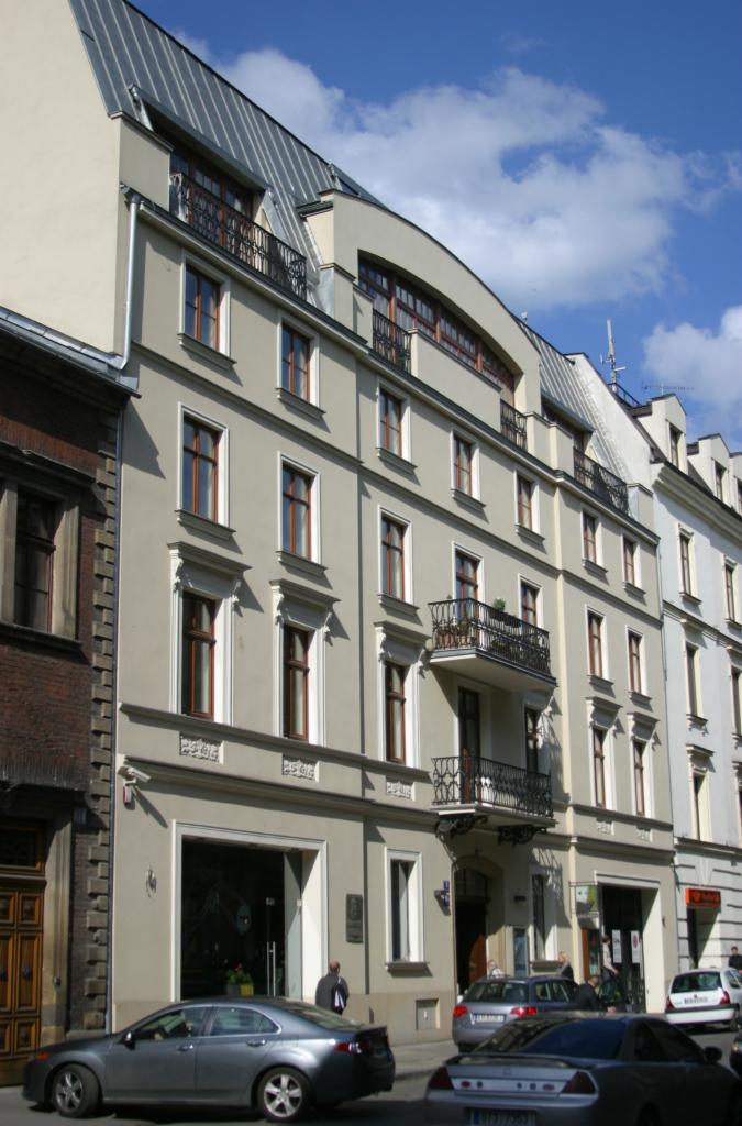 Odnowiona elewacja kamienicy w jasnych barwach z balkonami i metalowym dachem. Na dole widoczne zaparkowane samochody i fragment chodnika.